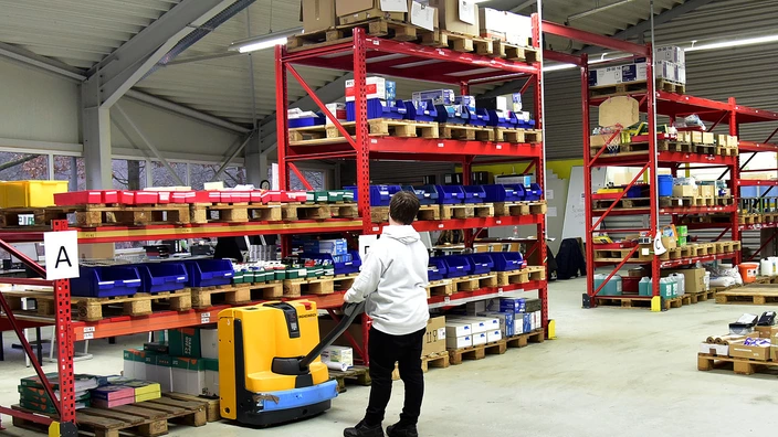 Ein Auszubildender fährt mit einem Hubwagen eine Palette aus einem Regal