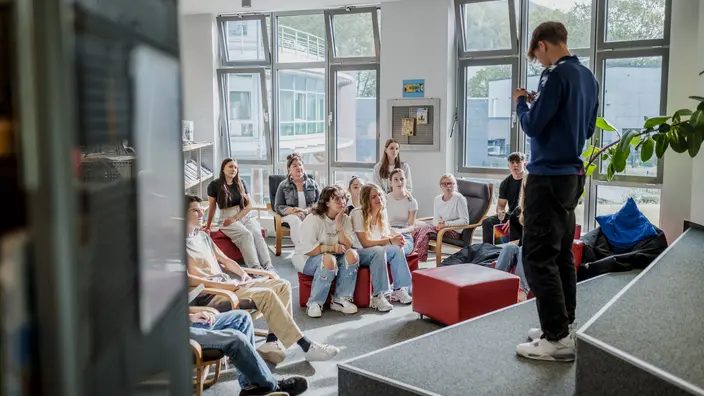 Ein Schüler steht auf einer Art Bühne vor einer kleinen Gruppe von Schülern und Schülerinnen und hält einen Vortrag.
