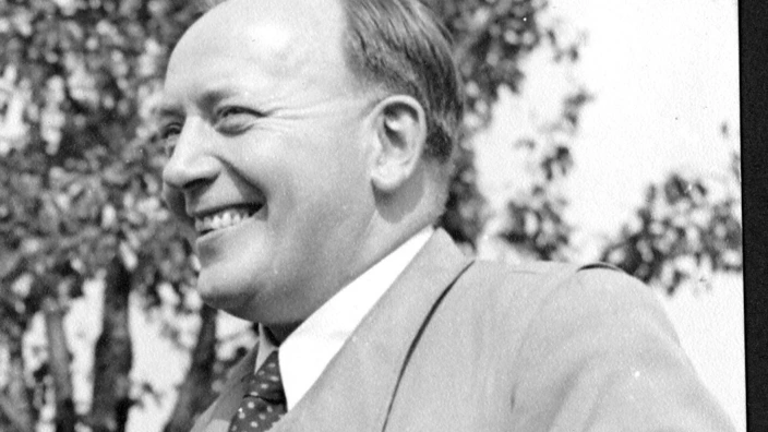 Schwarzweißfoto: Arnold Dannenmann im mittleren Alter mit zurückgekämmten Haaren steht im Freien und lächelt. Er trägt einen doppelreihigen Mantel mit Gürtel und Krawatte. Im Hintergrund ist ein Baum mit belaubten Ästen zu erkennen. Die Aufnahme wirkt historisch, vermutlich aus der Mitte des 20. Jahrhunderts. Der Mann wirkt freundlich und in ein Gespräch vertieft.