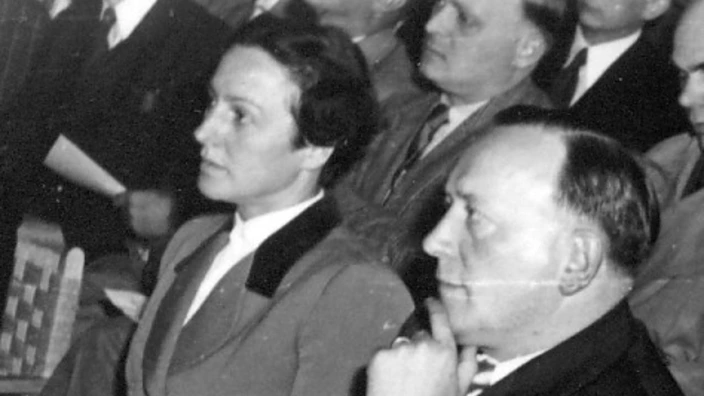  Schwarz/weis Ablichtung von Arnold Dannenmann mit seiner Frau Käthe 1952 zu Besuch im Jugenddorf Elze. Im Hintergrund sind mehrere Menschen vermutlich Lehrer zu sehen. Alle sitzen auf Holzstühlen und schauen Interessiert nach vorn. Es hat den Anschein als würden sie einem Redner gespannt zuhören. Arnold hat den Daumen der rechten Hand unterm Kinn und den Zeigefinger selbiger Hand am Kinn. Die Hände seiner Frau liegen gefaltet auf ihrem Schoß.