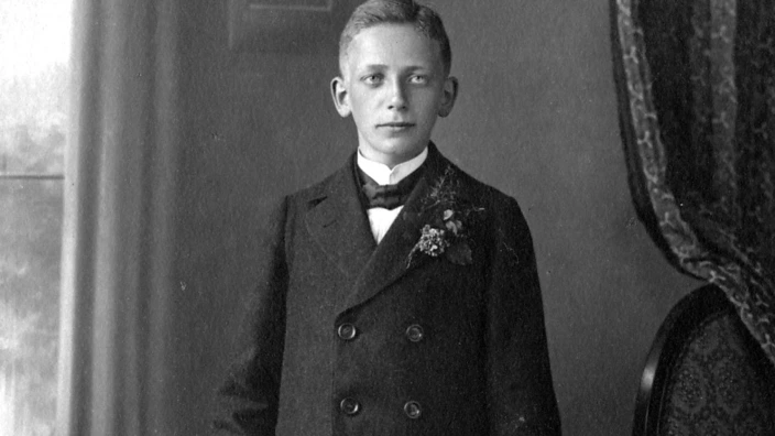 Arnold Dannemann im Anzug mit Blumengesteck zu seiner Konfirmation. In seiner linken Hand eine Bibel. Seine rechte Hand ruht auf einem kleinen Beistelltisch auf dem auch ein zum Anzug passender Hut liegt. Am Kragen seines Anzugs findet sich ein Blumengesteck.