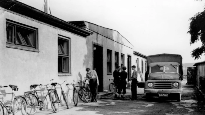 Schwarzweißfoto: Vor einem langgestreckten, einfachen Gebäude mit mehreren Fenstern stehen mehrere Fahrräder. Vier Männer unterhalten sich am Eingang, während ein älteres Nutzfahrzeug mit offener Ladefläche und der Aufschrift "S-AY 662" davor parkt. Die Szene wirkt wie ein typischer Arbeitsalltag in den 1950er- oder 1960er-Jahren, vermutlich in einem Werkstatt- oder Lagerumfeld.