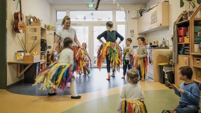 Pädagoginnen und Kinder tanzen in Röcken aus bunten Bändern. Ihre Spende schenkt Kindern in Kindergärten Geborgenheit.
