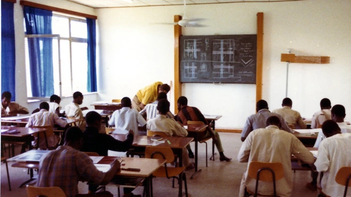 Farbfoto eines Klassenzimmers, vermutlich aus den 1970er- oder 1980er-Jahren: In einem hellen Raum mit Tafel und großen Fenstern sitzen zahlreiche Schüler in Reihen an Einzeltischen. Eine Lehrkraft steht an einem der Tische und hilft einem Schüler. Auf der Tafel sind technische Zeichnungen zu sehen – vermutlich handelt es sich um einen Unterricht in Mathematik oder Technik.