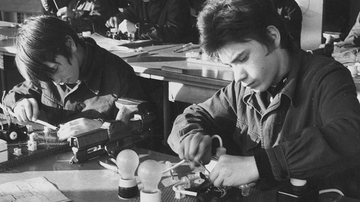 Schwarz-Weiß-Foto aus den 1970er-Jahren: Jugendliche arbeiten konzentriert in einem Werkraum an elektrischen Schaltungen. Zwei Jungen im Vordergrund schrauben an Versuchsplatten mit Glühbirnen und Schaltern. Im Hintergrund sitzen weitere Schüler an Werkbänken.