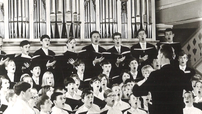 Schwarz-Weiß-Foto eines Chorkonzerts, Altensteiger Kirche 1962: Eine große Gruppe von Jungen und Mädchen in weißer Kleidung singt unter der Leitung eines Dirigenten. Im Hintergrund ist eine große Kirchenorgel zu sehen. Der Chor ist in mehreren Reihen angeordnet und hält Notenmappen in den Händen. Die Sänger:innen blicken konzentriert nach vorne, viele mit geöffnetem Mund im Gesang.