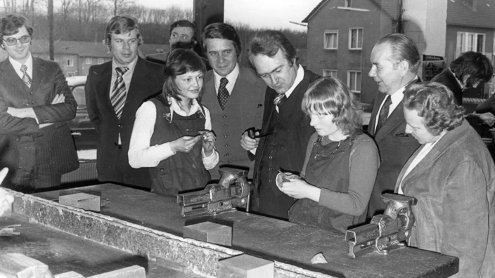 Schwarz-Weiß-Foto eines Werkraumbesuchs in den 1970er Jahren: Zwei junge Frauen in Arbeitskleidung arbeiten an Schraubstöcken, während eine Gruppe von Männern und Frauen – vermutlich Besucher:innen oder Offizielle – ihnen interessiert über die Schulter schaut. Die Szene spielt in einem Werkraum mit großen Fenstern und typischer Ausstattung.