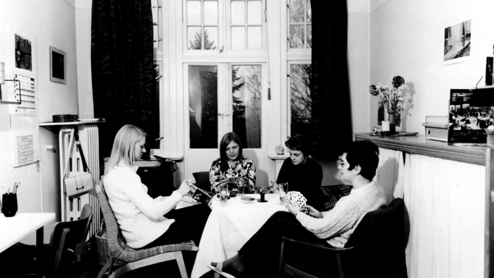 Schwarz-Weiß-Foto einer gemütlichen Teerunde in einem Wohnraum (vermutlich 1960er/1970er Jahre): Vier junge Frauen sitzen um einen kleinen Tisch mit Tischdecke, Getränken und Tellern. Sie unterhalten sich entspannt. Der Raum hat hohe Fenster mit dunklen Vorhängen und ist schlicht eingerichtet mit Stühlen, einem Schrank, Bildern und Pflanzen.