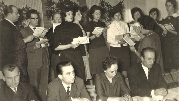 Schwarz-Weiß-Foto einer Chorprobe oder festlichen Veranstaltung (vermutlich 1950er–1960er Jahre): In der oberen Bildhälfte stehen elf Personen, die Notenblätter oder Liedtexte in den Händen halten und singen. In der unteren Bildhälfte sitzen sieben Personen an einem Tisch, offenbar in ein Programm oder eine Veranstaltung eingebunden. Die Szene wirkt formell, möglicherweise eine Feier oder ein offizieller Anlass.