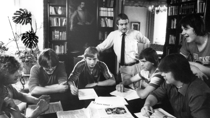 Schwarz-Weiß-Foto einer Gruppenszene bei der Zeitungsarbeit (vermutlich 1970er oder 1980er Jahre): Sieben junge Männer sitzen um einen Tisch, der mit Zeitungen und Schreibmaterialien bedeckt ist. Ein älterer Mann steht in der Mitte im Hintergrund, offenbar als betreuende oder leitende Person. Die Atmosphäre wirkt konzentriert und lebendig. Im Raum stehen Regale mit Büchern, im Hintergrund ist ein Gemälde zu sehen.