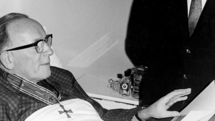 Schwarz-Weiß-Foto: Ein älterer Mann (Arnold Dannenmann) liegt in einem Krankenhausbett und trägt ein großes Kreuz (Bundesverdienstkreuz) um den Hals. Er schaut lächelnd auf einen Mann im Anzug, der neben dem Bett steht und mit ihm spricht. Die Szene wirkt feierlich und würdevoll.