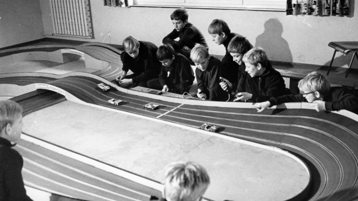 Schwarz-Weiß-Foto aus den 1960er-Jahren: Eine Gruppe von Jungen sitzt konzentriert um eine große Carrera-Rennbahn. Mit Handreglern steuern sie die kleinen Modellautos, die über die mehrfach geschwungene Bahn fahren. Die Szene spielt sich in einem einfachen Raum mit großem Fenster im Hintergrund ab.