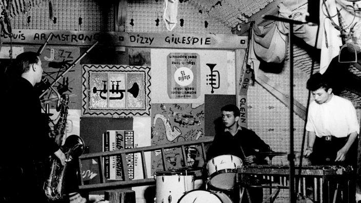 Schwarz-Weiß-Foto einer Jazzband in einem kreativ gestalteten Proberaum (vermutlich 1950er–1960er Jahre): Drei junge Männer musizieren – einer spielt Saxofon, einer sitzt am Schlagzeug, ein dritter am Vibrafon. Die Wände sind bunt dekoriert mit Jazz-Postern, Schallplatten und den Namen berühmter Musiker wie Louis Armstrong, Dizzy Gillespie und Gene Krupa. Die Atmosphäre wirkt künstlerisch und enthusiastisch.