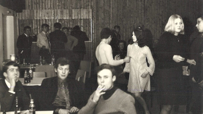 Schwarz-Weiß-Foto einer Abendveranstaltung (vermutlich 1960er–1970er Jahre): Im Vordergrund sitzen vier junge Männer an Tischen, einige mit Getränkeflaschen, einer raucht. Im Hintergrund tanzen mehrere junge Menschen, während andere in Gruppen stehen und sich unterhalten. Die Szene wirkt ungezwungen und gesellig, möglicherweise eine Feier oder ein Tanzabend.