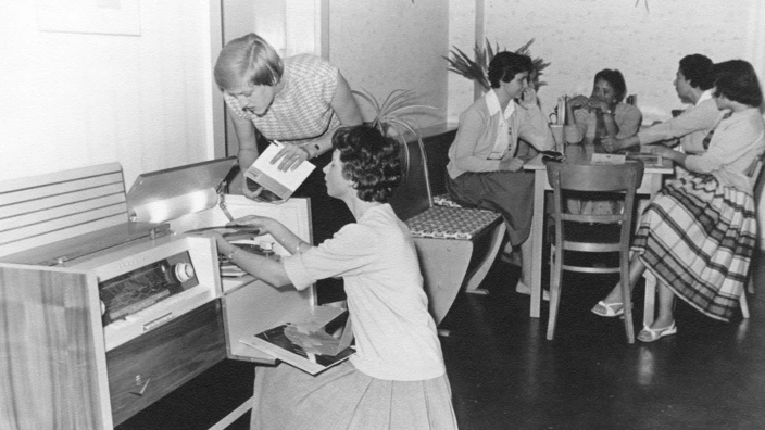 Zwei Frauen bedienen ein Schallplattenradio, eine legt eine Schallplatte auf, während die andere ein Cover betrachtet. Im Hintergrund sitzen mehrere Frauen um einen Tisch, vertieft in Gespräche. Die Szene wirkt wie ein geselliges Beisammensein in den 1950er- oder 60er-Jahren.