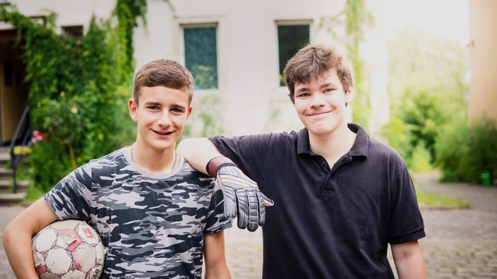 Zwei Jugendliche stehen nebeneinander und lächeln in die Kamera. Ein junger Mann hält einen Fußball unter dem Arm. Der andere junge Mann legt seinen Arm auf der Schulter des anderen ab.