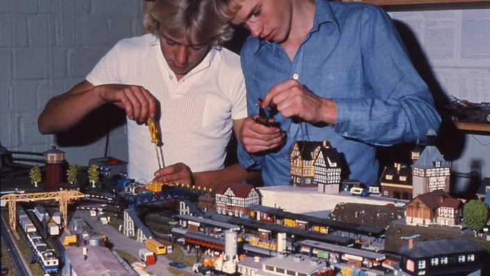Farbfoto: Zwei Jugendliche arbeiten konzentriert an einer großen Modelleisenbahnanlage. Einer nutzt einen Schraubenzieher, der andere hält eine Figur. Vor ihnen eine detailreiche Miniaturlandschaft mit Zügen, Gleisen, Häusern und Fahrzeugen. Die Szene vermittelt handwerkliches Geschick und Leidenschaft fürs Basteln.