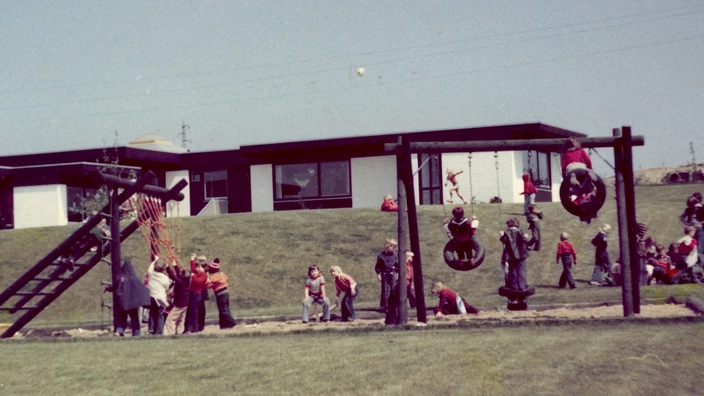 Farbfoto von 1975: Kinder spielen auf einem großzügigen Außengelände mit moderner Kletter- und Schaukellandschaft vor einem flachen Kita-Gebäude. Einige Kinder klettern am Netz, andere schaukeln in Reifen oder sitzen auf dem Rasen. Die Szene zeigt die erste Kindertagesstätte im CJD Extertal bei sonnigem Wetter.
