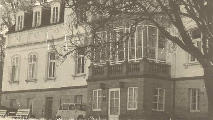 Sepiafarbenes Foto eines historischen Schlossgebäudes mit großen Fenstern, Mansarddach und Wintergarten-Erker. Vor dem Gebäude neben der Tür mit CJD-Logo steht ein älteres Fahrzeug, daneben zwei Parkbänke. Ein kahler Baum ragt dominant ins Bild, im Vordergrund eine Wiese. Die Szenhig und zeittypisch für die Mitte des 20. Jahrhe wirkt ruunderts.