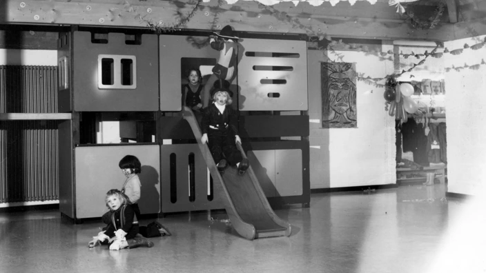 Schwarz-weiß-Foto einer geschmückten Kita-Spielhalle: Fünf Kinder spielen an einem großen Klettergerüst mit Rutsche. Ein Kind im Kostüm rutscht gerade hinunter, andere stehen oder sitzen spielend davor. Girlanden und Luftballons deuten auf eine Feier hin. Die Atmosphäre ist fröhlich und lebendig.