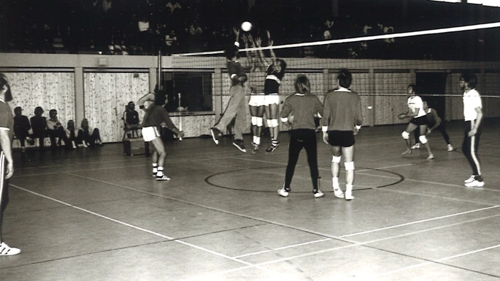 Schwarz-weiß-Foto aus einer Sporthalle im Jahr 1974: Zwei Volleyballteams stehen sich bei einem Spiel gegenüber. Drei Spieler springen am Netz zum Block oder Angriff hoch, der Ball schwebt darüber. Zuschauerinnen und Zuschauer sitzen am Rand der Halle, die Atmosphäre wirkt konzentriert und sportlich. Aufgenommen während der Sportwoche am CJD Altensteig.