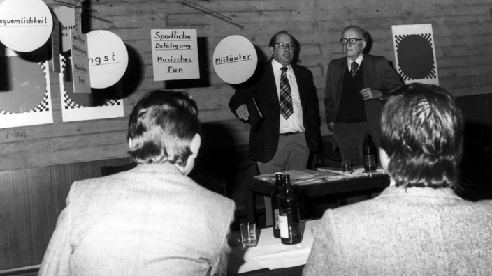 Schwarz-weiß-Foto von 1978: Zwei Männer stehen bei einer pädagogischen Veranstaltung in Bad Teinach vor einer kleinen Gruppe Zuhörer. An der Wand hängen Plakate mit Begriffen wie „Angst“, „Bequemlichkeit“, „Mitläufer“ und „Sportliche Betätigung“. Auf dem Tisch stehen Flaschen und Gläser, die Stimmung wirkt konzentriert und sachlich.