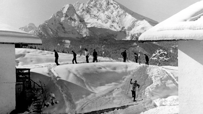Schwarz-weiß-Foto aus den 1970er Jahren: Eine Gruppe junger Menschen auf Langlaufskiern bewegt sich über eine verschneite Anhöhe zwischen zwei Gebäuden. Im Hintergrund erhebt sich das imposante Watzmann-Massiv über den schneebedeckten Wald. Die Szene wirkt ruhig und konzentriert, typisch für eine Wintersportübung im CJD Berchtesgaden.