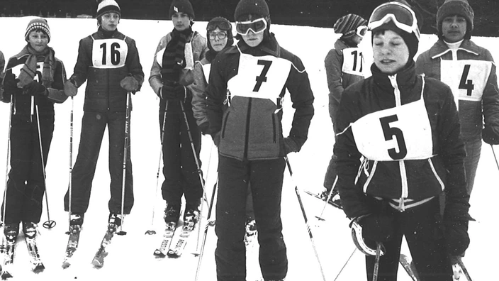 Schwarz-weiß-Foto einer Skifreizeit des CJD Altensteig in den 1970er Jahren. Rund 30 Personen in Skikleidung posieren lachend vor einer winterlichen Berglandschaft. Einige halten Skier, andere stehen locker in Gruppen. Im Hintergrund verschneite Hänge und Bäume. Kleidung und Ausrüstung sind typisch für die 70er Jahre und vermitteln Gemeinschaft und Freude im Schnee.