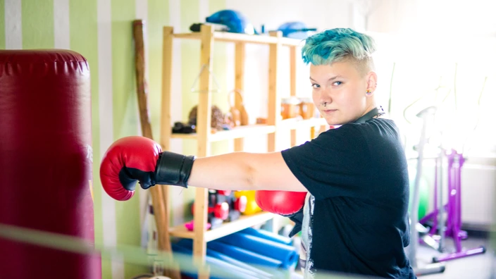 Eine Personen mit blauen Haaren und roten Boxhandschuhen steht vor einem Boxsack.