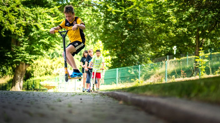 Ein Kind springt mit einem Roller in die Luft. Im Hintergrund stehen drei andere Kinder mit ihren Rollern. 