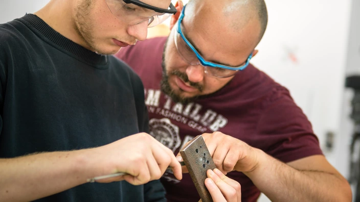 Zwei Personen mit Arbeitsschutzbrillen schauen sich ein Metallstück an.