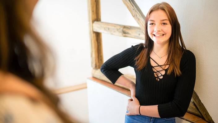 Eine junge Frau lehnt an eine Wand und unterhält sich mit einer Person, die ihr gegenüber steht.