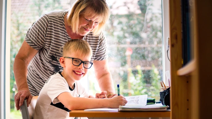 Ein Kind sitzt am Schreibtisch und macht Hausaufgaben. Eine Pädagogin steht hinter ihm und schaut in sein Heft.