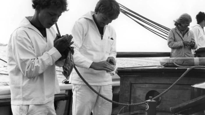 Schwarz-weiß-Foto an Bord eines Segelschiffs: Zwei Jugendliche in heller Arbeitskleidung polieren ein Seil mit Tuch und Politur. Im Hintergrund arbeiten zwei weitere Personen auf dem Deck. Das Meer ist ruhig, Takelage und Holzaufbauten deuten auf ein traditionelles Segelschiff hin. Die Szene wirkt konzentriert und vermittelt Teamarbeit und maritimes Handwerk.
