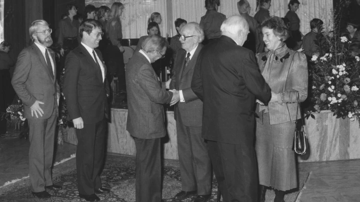 Schwarz-weiß-Foto einer feierlichen Gratulationsszene: Mehrere Personen stehen auf einem Teppich und reichen einem älteren Herrn nacheinander die Hand – vermutlich zu seinem 80. Geburtstag. Im Hintergrund ein Chor junger Menschen auf einer Bühne, Blumenschmuck rechts im Bild. Die Atmosphäre ist festlich und respektvoll.