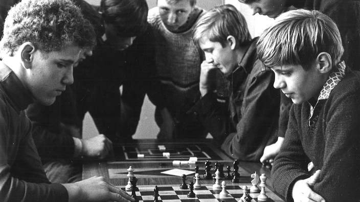 Schwarz-weiß-Foto von etwa 1969: Zwei Jungen spielen konzentriert Schach, mehrere weitere Jugendliche beugen sich interessiert über das Brett. Die Partie scheint anspruchsvoll, die Atmosphäre ist still und fokussiert. Im Hintergrund ist ein weiteres Brettspiel zu erkennen. Das Bild vermittelt Nachdenklichkeit, Strategie und gemeinsames Lernen in ruhiger Umgebung.