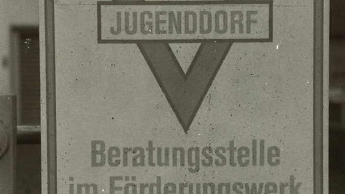 Schwarz-weiß-Foto eines alten Hinweisschilds am Gittertor. In der Mitte das CJD-Dreieck mit der Aufschrift „Jugenddorf“. Darunter steht: „Beratungsstelle im Förderungswerk für Spitzenbegabte“. Am unteren Rand klein: „Christliches Jugenddorfwerk Deutschlands“. Das Schild wirkt wettergezeichnet und stammt erkennbar aus der Zeit um 1989.