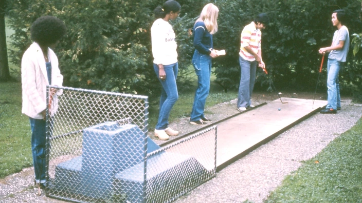Farbfoto aus den 1980er Jahren: Fünf junge Menschen spielen gemeinsam eine Runde Minigolf im Freien. Eine Person schlägt gerade den Ball, die anderen schauen zu. Die Szene spielt sich auf einer schmalen Betonbahn ab, umgeben von dichtem Grün. Die Kleidung und Frisuren sind typisch für die Zeit, die Stimmung wirkt ruhig und konzentriert.