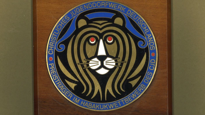 Farbfoto einer rechteckigen Holzplakette mit rundem Emblem in der Mitte: Ein stilisierter Löwenkopf mit roter Iris und goldener Mähne auf schwarzem Grund. Umgeben wird das Motiv von einem blauen Ring mit der goldenen Aufschrift „Christliches Jugenddorfwerk Deutschlands – Preisträger im Habakukwettbewerb des CJD“. Die Darstellung wirkt feierlich und symbolträchtig.