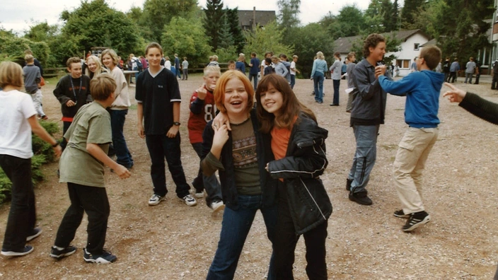 Farbfoto aus den 1990er Jahren: Eine Gruppe Jugendlicher steht und spielt auf einem Schulhof oder Gelände mit Kiesboden. Im Vordergrund lachen zwei Mädchen in die Kamera, Arm in Arm. Um sie herum unterhalten sich weitere Jugendliche, einige sind in Bewegung. Im Hintergrund Bäume, Häuser und ein lockeres, fröhliches Miteinander.