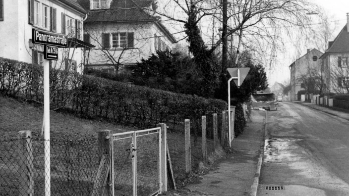Schwarz-weiß-Foto einer ruhigen Wohnstraße mit dem Straßenschild „Panoramastraße“. Links im Bild ein Gartenzaun mit Tor, dahinter Wohnhäuser mit Fensterläden. Rechts verläuft ein Gehweg mit einer Vorfahrtstafel, die Straße zieht sich in die Tiefe des Bildes. Winterliche Stimmung mit kahlen Bäumen und leichten Schneeresten am Straßenrand.