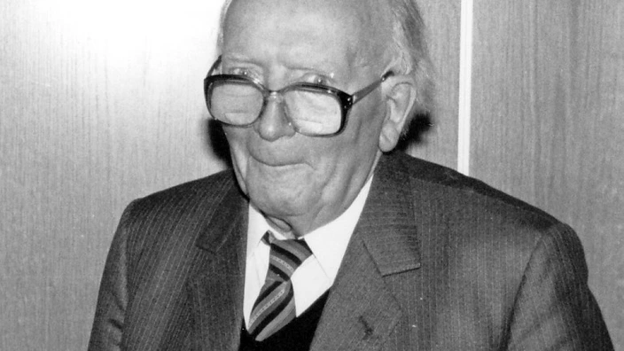 Schwarz-weiß-Porträt eines älteren Mannes mit Brille, Anzug und gestreifter Krawatte. Er steht vor einer Holzvertäfelung und lächelt leicht. Der Ausdruck wirkt freundlich und würdevoll. Das Bild stammt vermutlich aus den 1990er Jahren und zeigt den Mann in einem feierlichen oder offiziellen Moment.