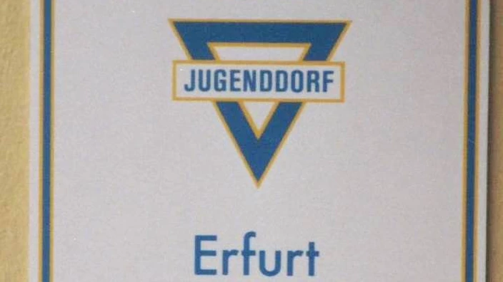 Farbfoto eines Hinweisschilds an einer Wand. Oben das Logo des CJD mit dem blauen Dreieck und dem Schriftzug „Jugenddorf“. Darunter steht: „CJD Erfurt Rehabilitationszentrum – Christliches Jugenddorfwerk Deutschlands e. V.“. Auf dem unteren Schild: „Geschützte Werkstätten, Havannaer Str. 29“. Das Schild stammt vermutlich aus den 1990er Jahren.
