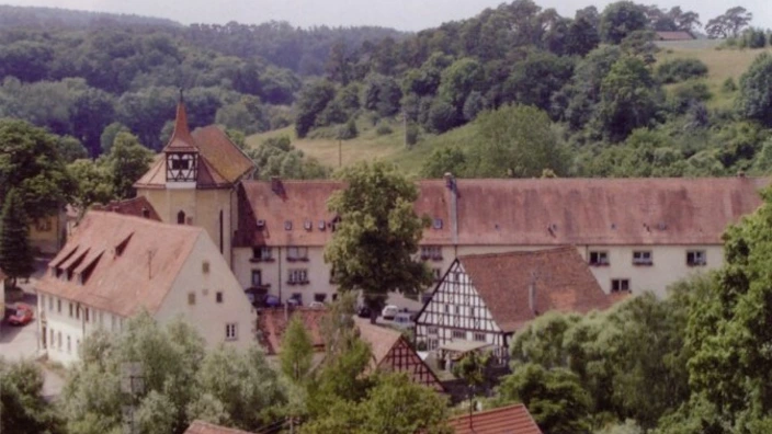 Farbfoto eines historischen Kloster- oder Gutshof-Ensembles mit roten Ziegeldächern und Fachwerkhäusern, eingebettet in eine grüne, hügelige Landschaft. Im Zentrum ein langgestrecktes Hauptgebäude mit Turm und Uhr. Im Vordergrund Bäume und Dächer, im Hintergrund dichter Wald. Die Szene wirkt ruhig und ländlich. Aufnahmeort: Projekt Chance im Kloster Frauenthal.