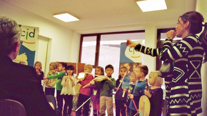 Farbfoto: Eine Gruppe von Kita-Kindern steht nebeneinander in einem Raum und führt mit bunten Instrumenten und selbstgebastelten Requisiten eine musikalische Darbietung auf. Eine Erzieherin dirigiert mit lebendiger Geste. Im Hintergrund zwei CJD-Rollups. Die Szene spielt sich offenbar bei einer feierlichen Eröffnung oder Veranstaltung in einer CJD-Kita in Berlin ab.