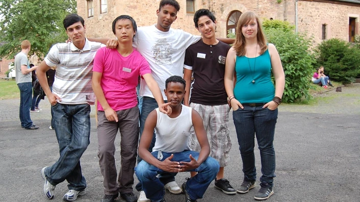 Farbfoto: Sechs junge Menschen stehen und knien lächelnd in einer lockeren Pose vor einem historischen Gebäude mit Sandsteinfassade. Sie tragen Namensschilder, was auf eine Veranstaltung oder ein Seminar hinweist. Im Hintergrund weitere Teilnehmende auf dem Gelände. Die Szene vermittelt Offenheit, Vielfalt und Gemeinschaft.