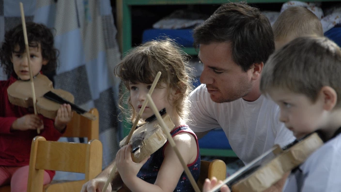 Farbfoto: Ein Erzieher unterstützt ein Mädchen beim Spielen einer selbstgebastelten Pappgeige. Weitere Kinder sitzen daneben mit ähnlichen Instrumenten und konzentrieren sich auf das Spiel. Die Szene spielt sich in einem Gruppenraum mit Deckenlagern im Hintergrund ab. Das Bild vermittelt kreative musikalische Frühförderung und liebevolle Begleitung im Kita-Alltag.