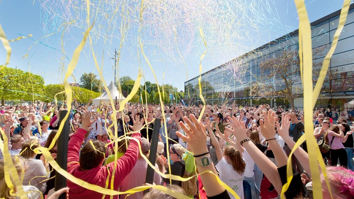 Farbfoto: Eine große Menschenmenge feiert bei sonnigem Wetter ein buntes Festival im Freien. Zahlreiche Personen strecken jubelnd die Arme in die Luft, während gelbe und bunte Luftschlangen durch die Luft wirbeln. Im Hintergrund sind Zelte, Bäume und ein modernes Gebäude mit Glasfassade zu sehen. Die Stimmung wirkt fröhlich und ausgelassen.