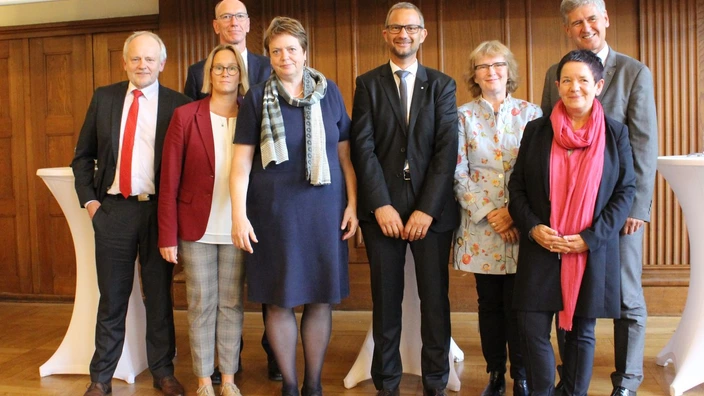 Farbfoto: Acht Personen stehen für ein Gruppenfoto in einem holzvertäfelten Saal aufgereiht. Alle sind formell gekleidet, teils in Anzug, Kostüm oder Blazer. Zwei Stehtische mit weißen Hussen flankieren die Gruppe. Die Stimmung wirkt professionell und freundlich. Es handelt sich vermutlich um eine offizielle Veranstaltung, Tagung oder Projektpräsentation im Bildungs- oder Sozialbereich.