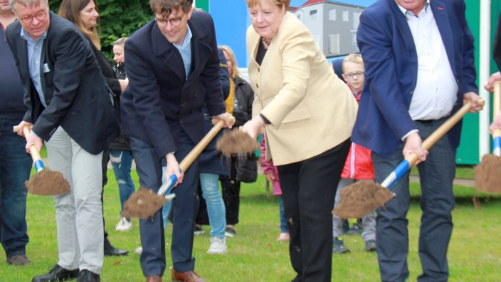 Farbfoto: Vier Personen in festlicher Kleidung führen gemeinsam einen symbolischen Spatenstich auf einer Wiese durch. In der Mitte Bundeskanzlerin a. D. Angela Merkel mit beigefarbener Jacke. Im Hintergrund ein Bauwagen mit Visualisierung eines geplanten Gebäudes und mehrere Zuschauer:innen. Die Szene markiert den feierlichen Start eines Bauprojekts mit politischer Beteiligung.